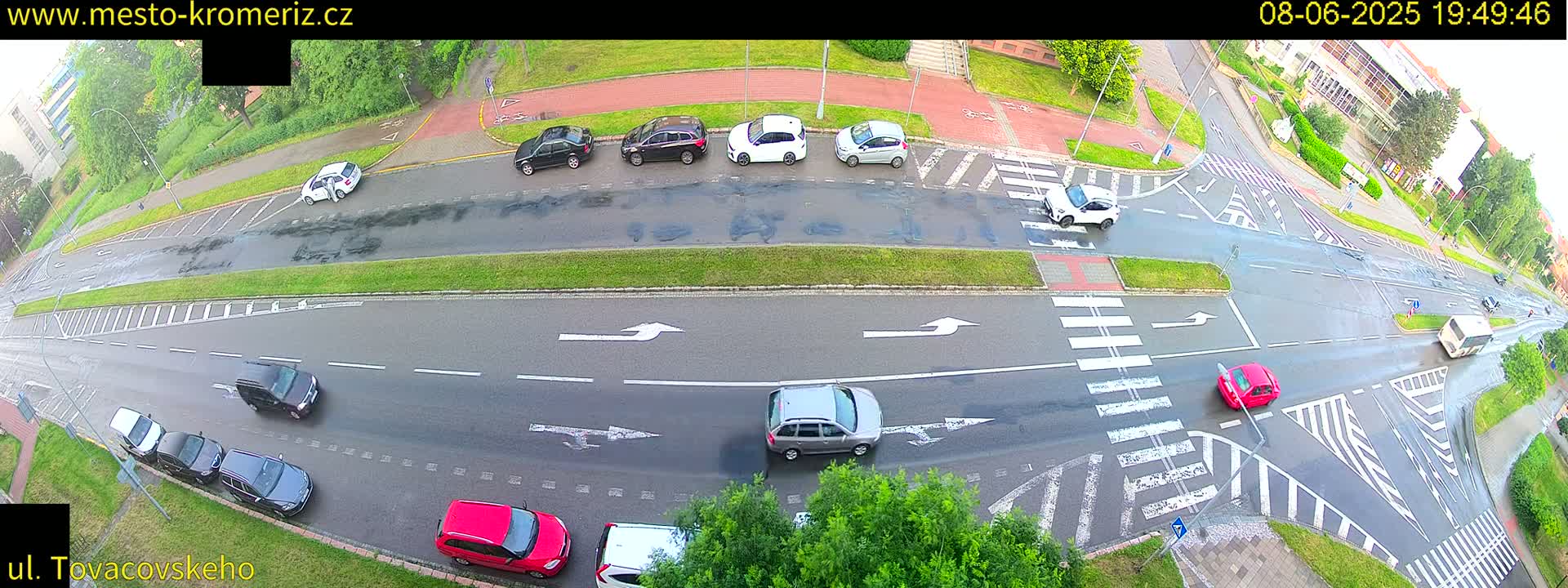 A wet road intersection with several cars, a pedestrian crossing, and green spaces surrounding it.