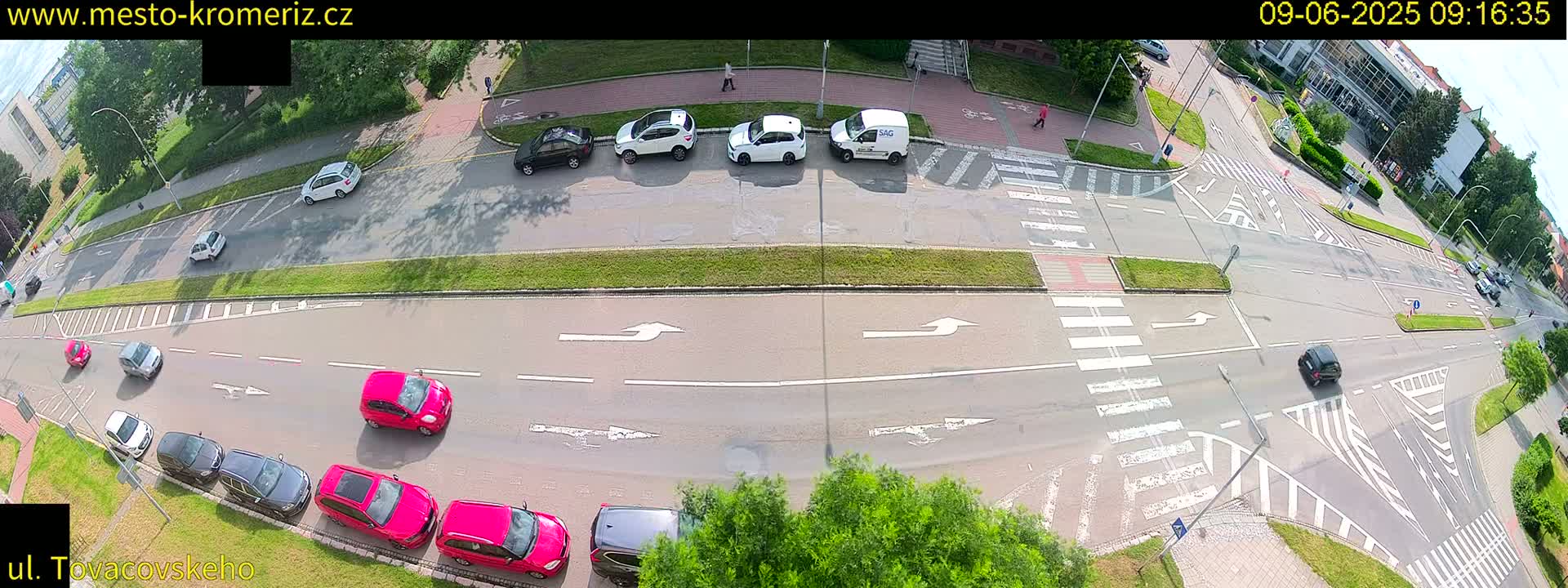 A high-angle view of a street intersection on a sunny day, showing several cars parked and driving on a multi-lane road with crosswalks and pedestrian paths.