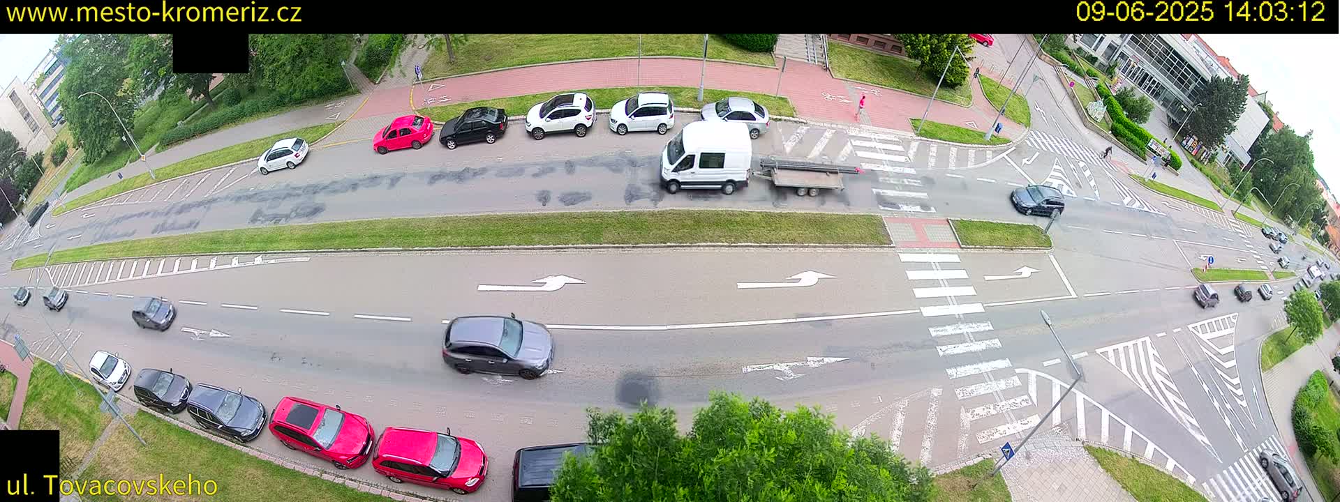 An aerial view shows several cars parked and driving on a roadway intersection on a partly sunny day.