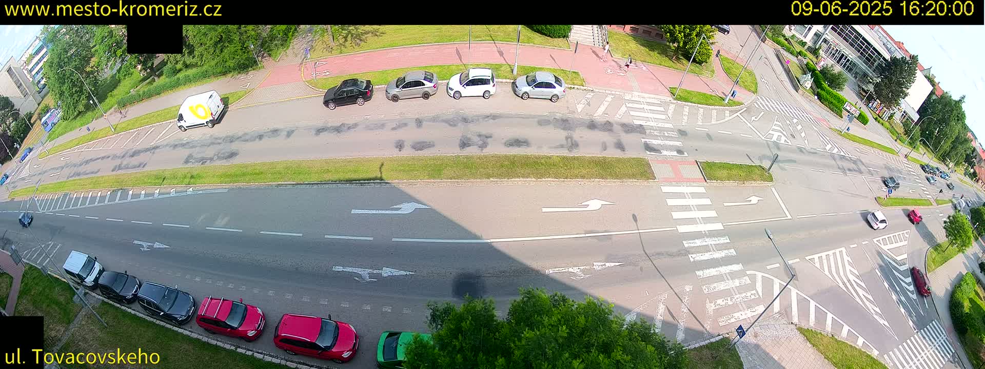 An overhead view of a sunny road intersection with several cars parked and driving, and pedestrians walking nearby.