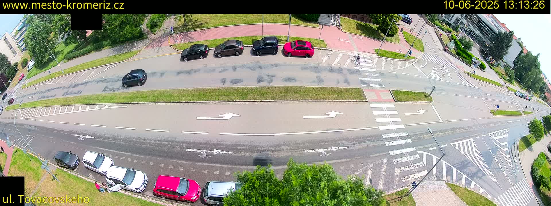 An aerial view of a road intersection on a sunny day shows several cars parked and driving, along with a cyclist crossing a crosswalk.