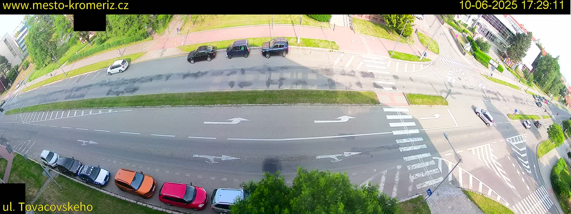 A high-angle, sunny view of a multi-lane road intersection with several cars parked and driving, featuring crosswalks and turn indicators painted on the asphalt.