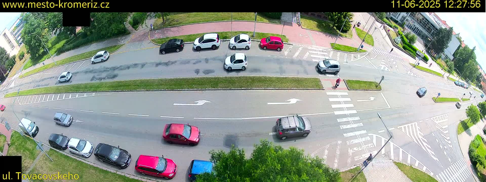 A high-angle view shows a sunny day with several cars driving and parked on a multi-lane road with crosswalks and bicycle lanes, surrounded by green landscaping.