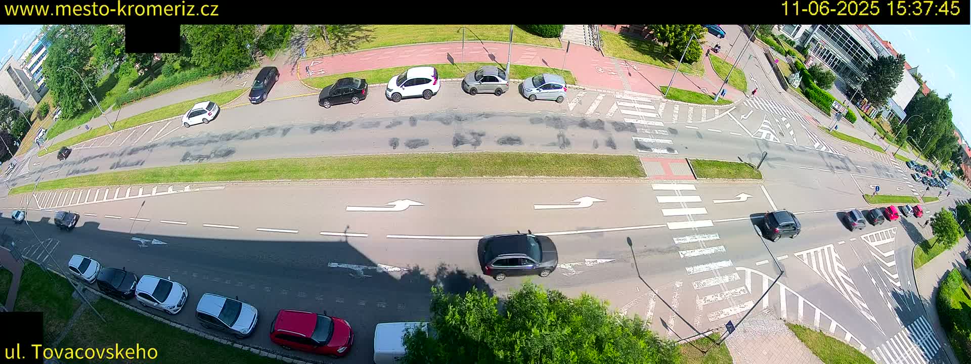 An overhead view of a multi-lane road intersection on a sunny day, showing several cars parked and driving, with pedestrian crosswalks and bicycle lanes.