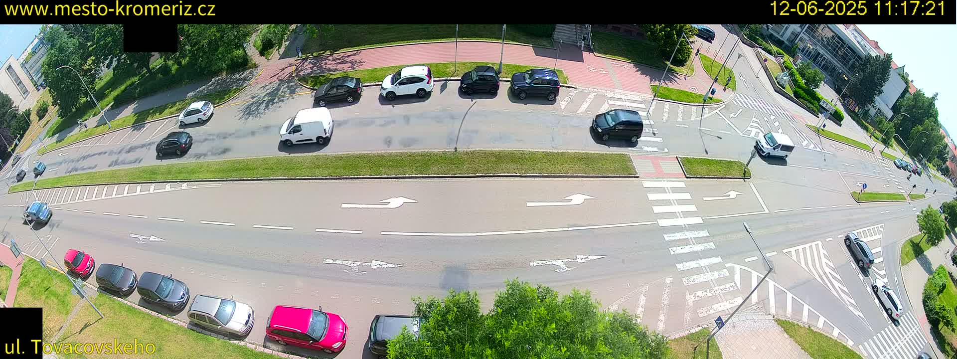 A sunny overhead view of a multi-lane road intersection with several cars and vans driving and parked, surrounded by green lawns and trees.