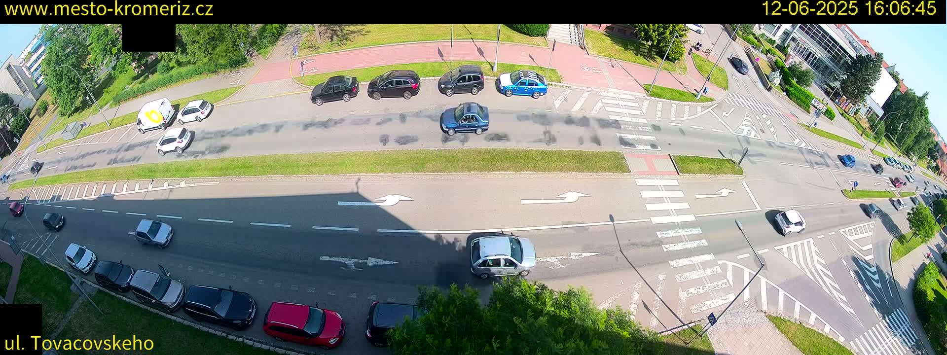 A high-angle view of a sunny street scene shows several cars driving on a multi-lane road with crosswalks and greenery alongside.