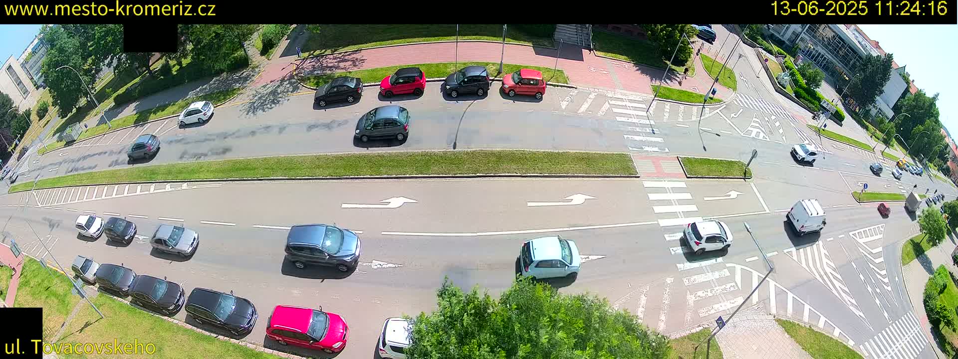 A high-angle view of a sunny city intersection shows several cars moving through the crosswalks and streets.
