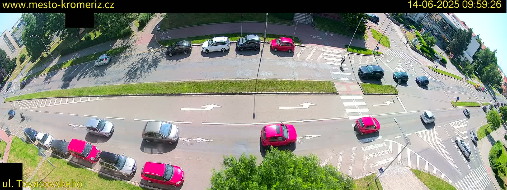 An aerial view shows a sunny day with numerous cars at a multi-lane intersection, some stopped at crosswalks and others moving through the intersection.