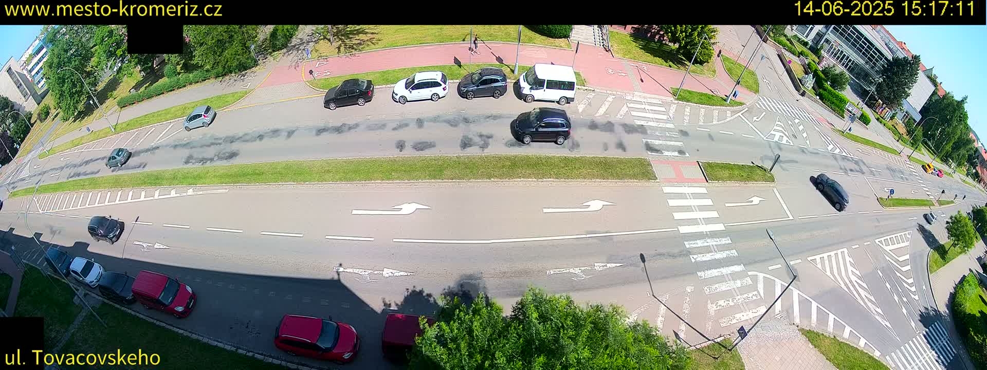 A sunny overhead view of a multi-lane road intersection with several cars and a van, including pedestrian crosswalks and bike lanes.