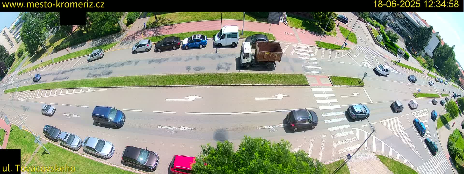 A sunny day reveals a bird's-eye view of a multi-lane road intersection with numerous cars and a truck, surrounded by green grass and buildings.