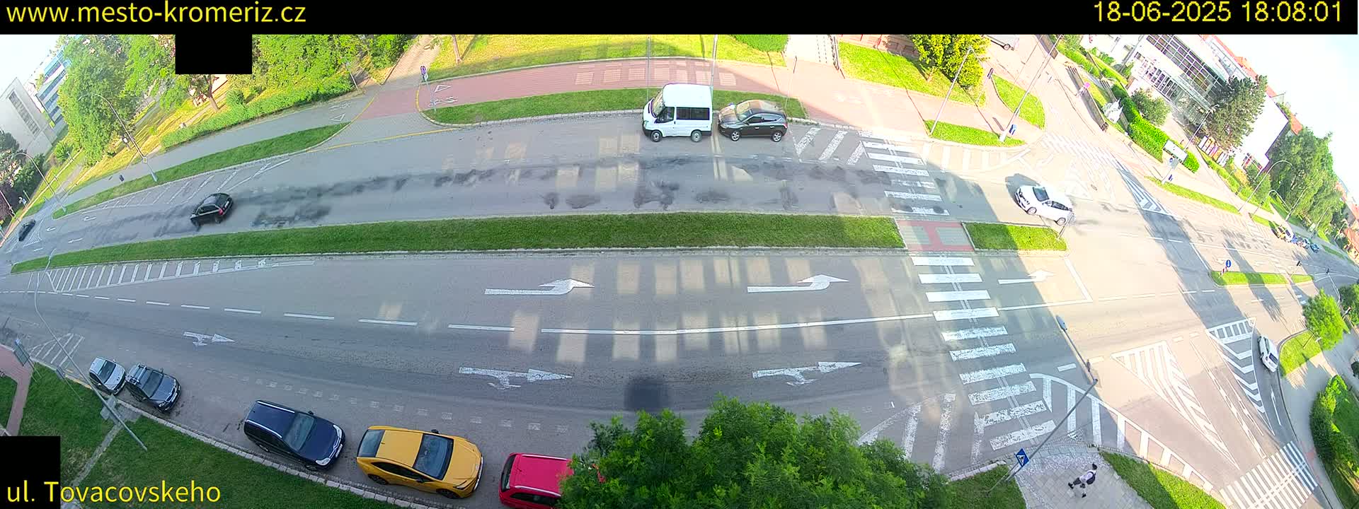 An aerial view of a street intersection on a sunny day shows several cars parked and driving on a multi-lane road with crosswalks and bicycle lanes.