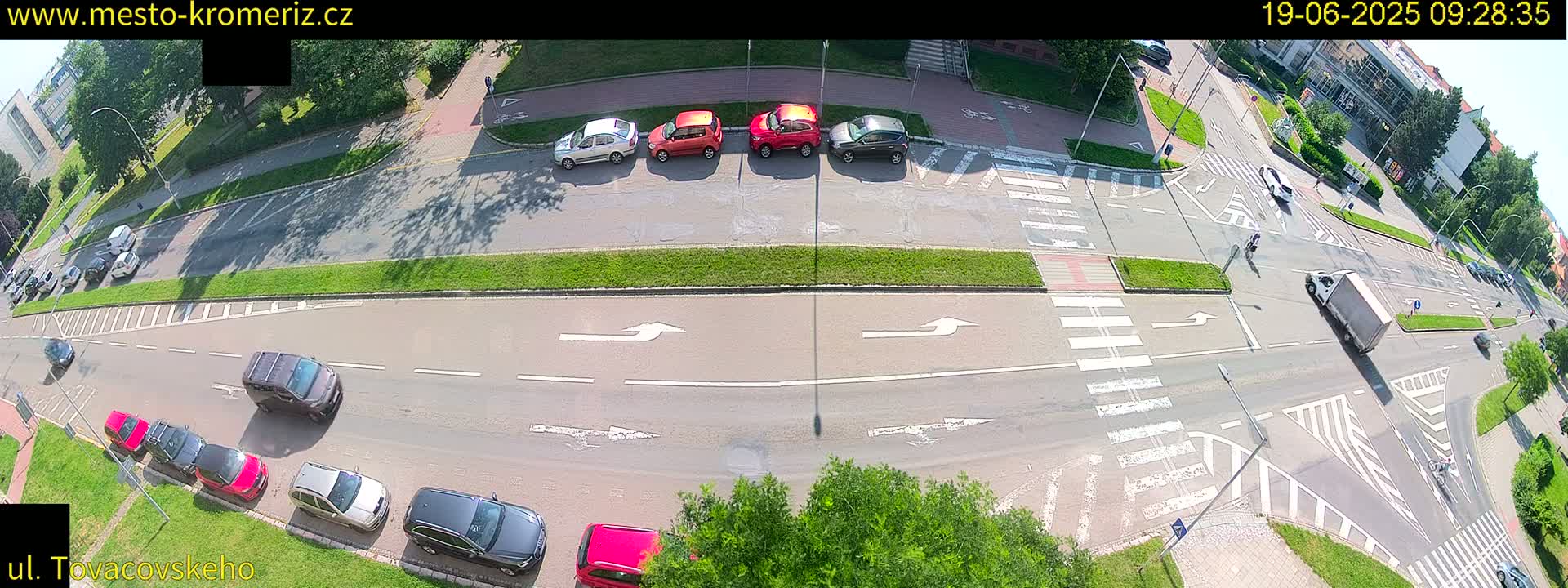 A high-angle, sunny day view of a multi-lane road intersection with several cars parked along the side and others driving through, with pedestrians and cyclists crossing.
