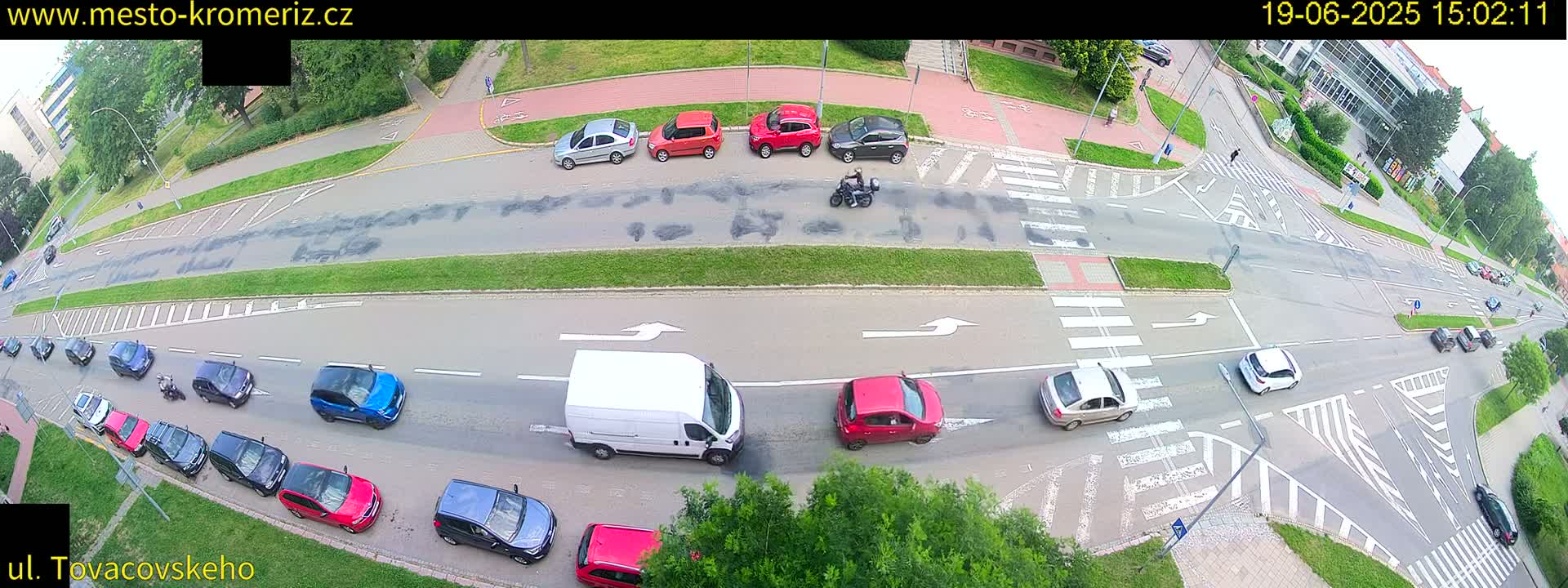 An aerial view of a road intersection with several cars and a motorcycle, under a partly sunny sky.