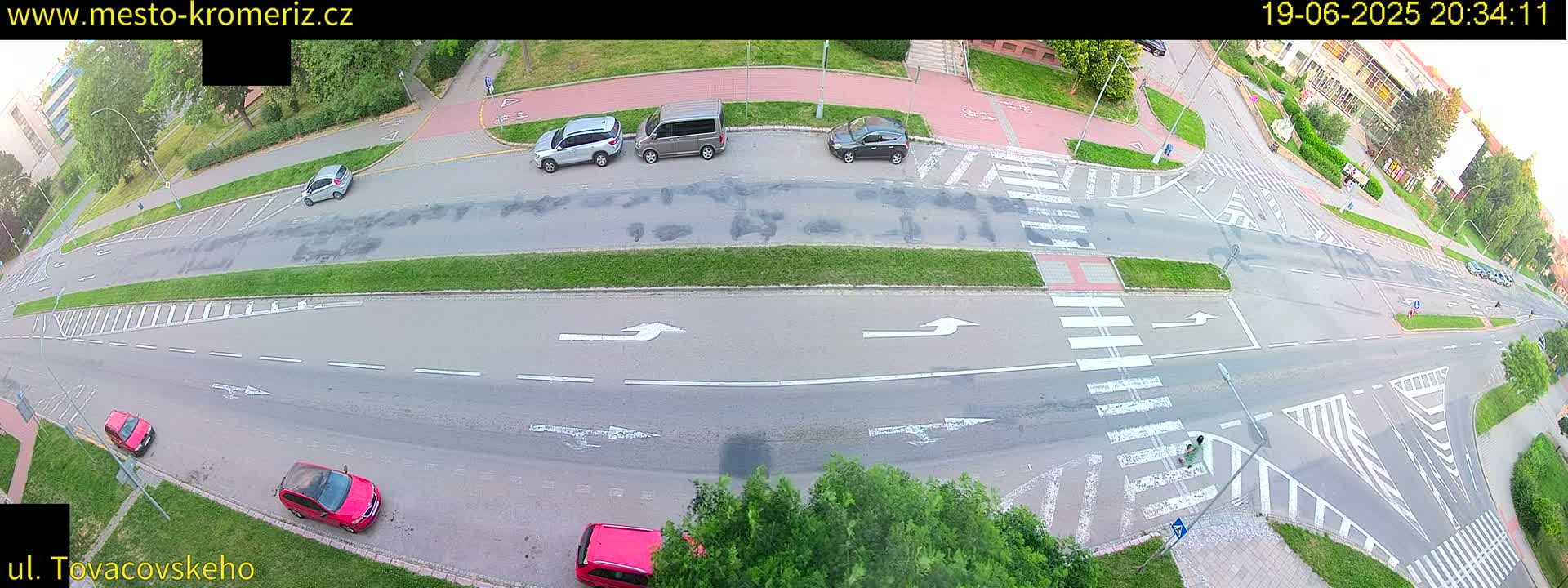An aerial view of a paved road intersection with several cars parked and driving, crosswalks, and surrounding green spaces under a partly cloudy sky.