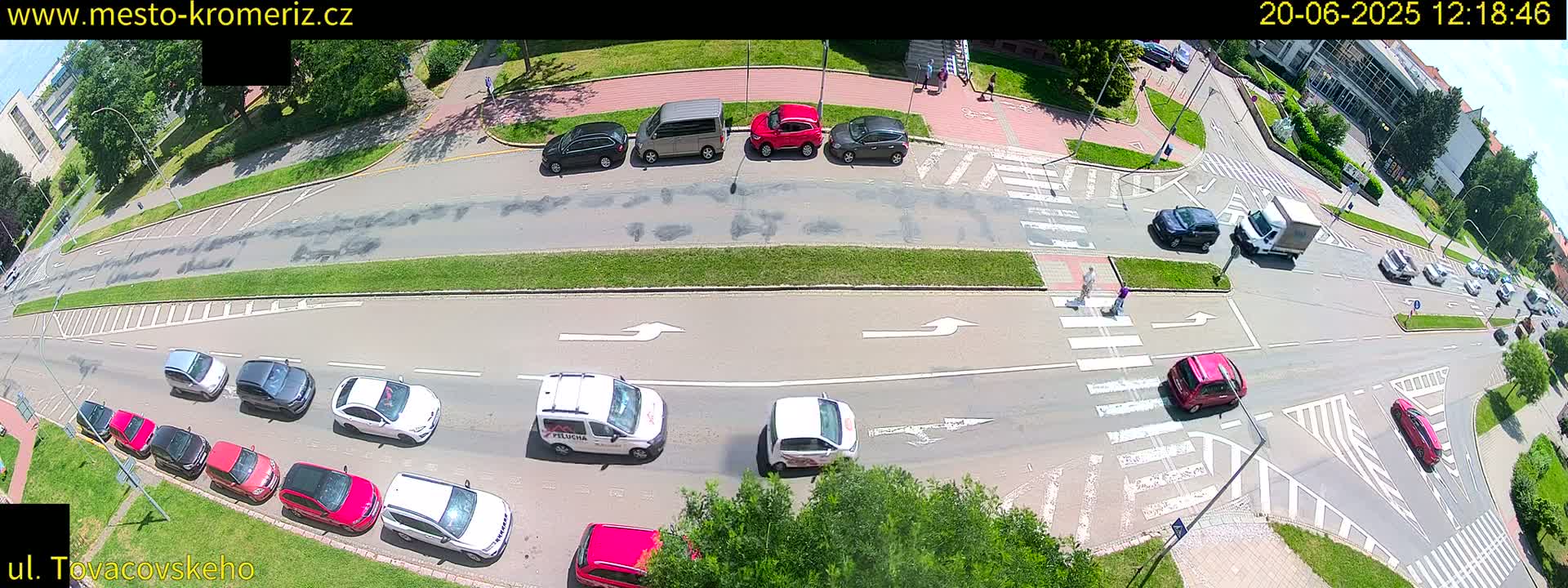 An aerial view of a sunny city intersection shows several cars stopped at crosswalks and traffic lights, with pedestrians crossing the street.