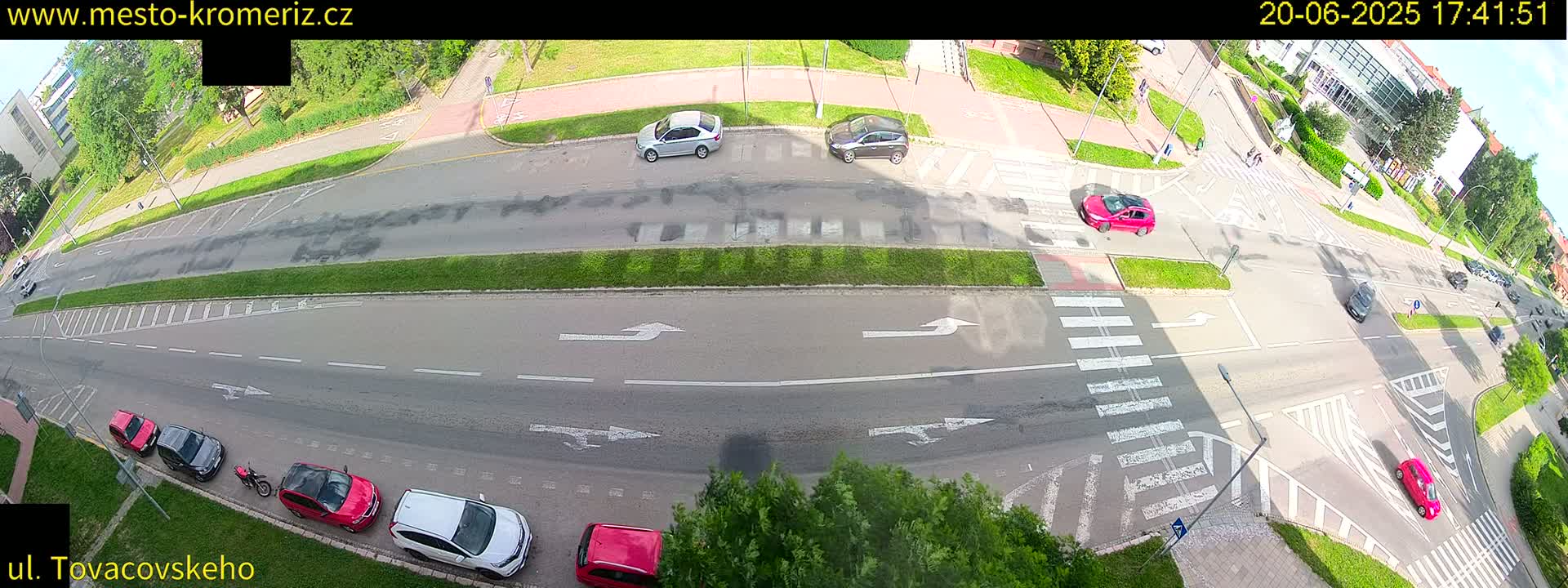A sunny overhead view of a multi-lane road intersection with several cars parked and driving, marked by crosswalks and turn arrows.