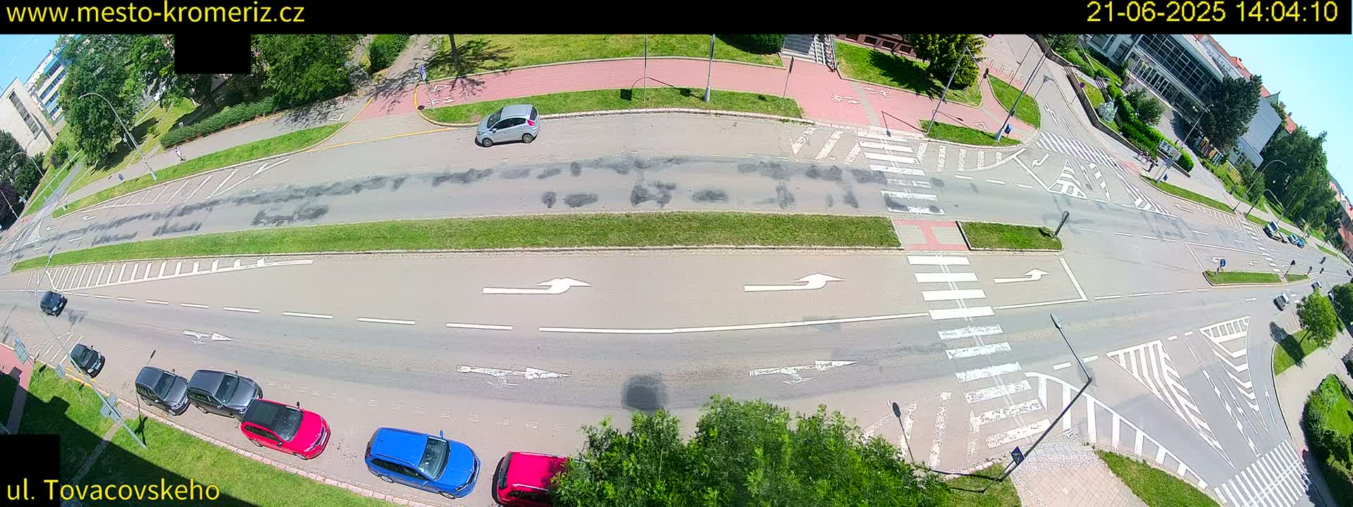 A high-angle view of a road intersection on a sunny day, showing several cars parked and driving on a multi-lane road with crosswalks and marked turning lanes.