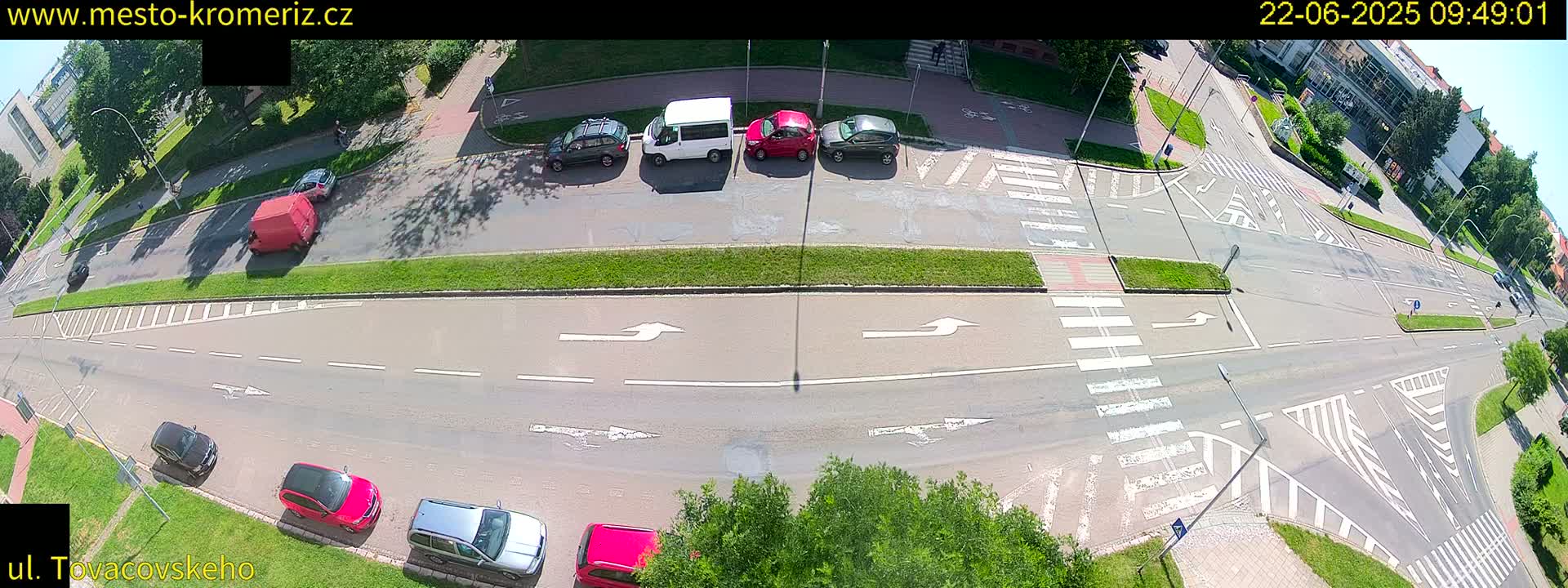 A sunny day overhead view of a street intersection with several cars parked and driving, marked by crosswalks and directional arrows.