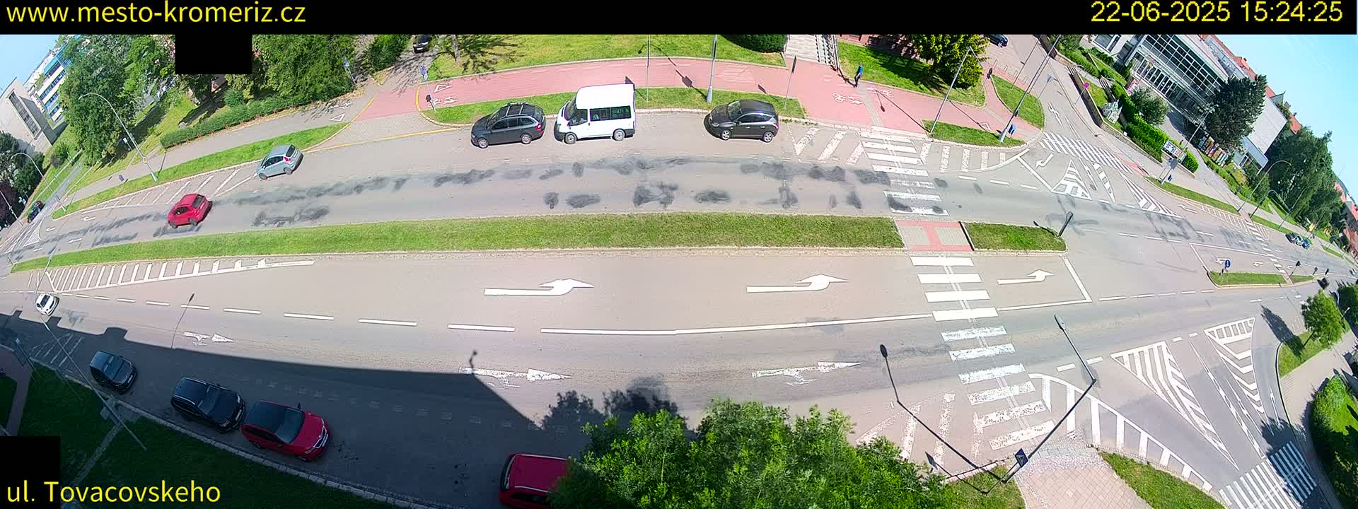 A sunny overhead view of a multi-lane road intersection with several cars and a van, marked with crosswalks and directional arrows.