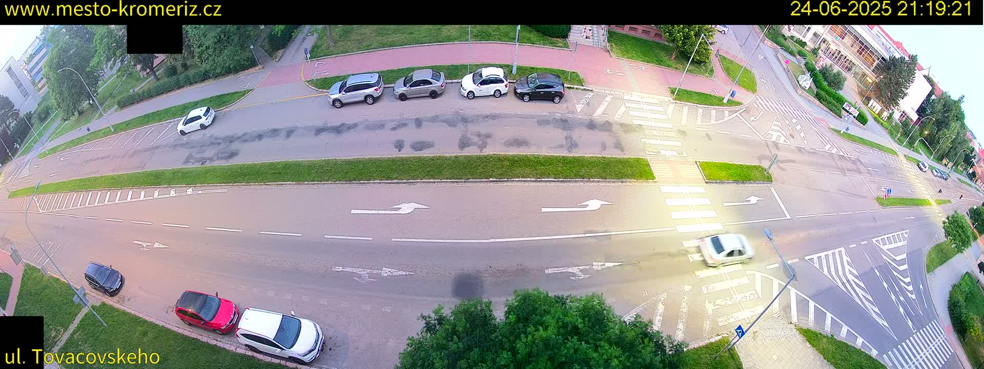 A high-angle, long shot of a lightly trafficked intersection at dusk, with several cars parked along the side of the road and a few driving through the intersection.