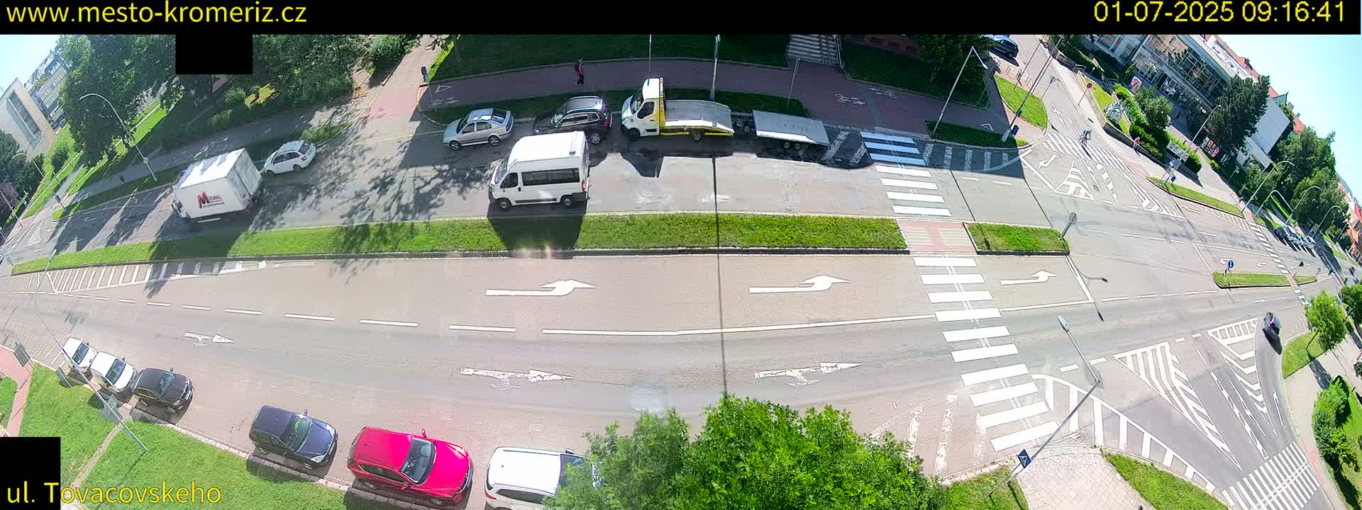 A sunny day shows a high-angle view of a road intersection with several cars, a van, and a tow truck, along with pedestrian crosswalks and surrounding greenery.