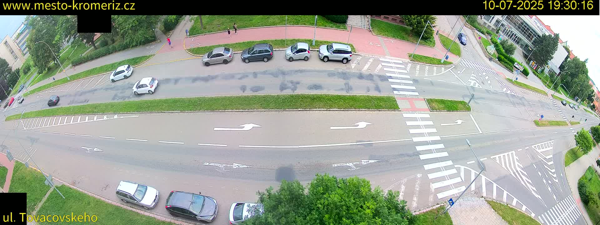 An overhead view of a roadway intersection with several cars and pedestrians, on a partly sunny day.