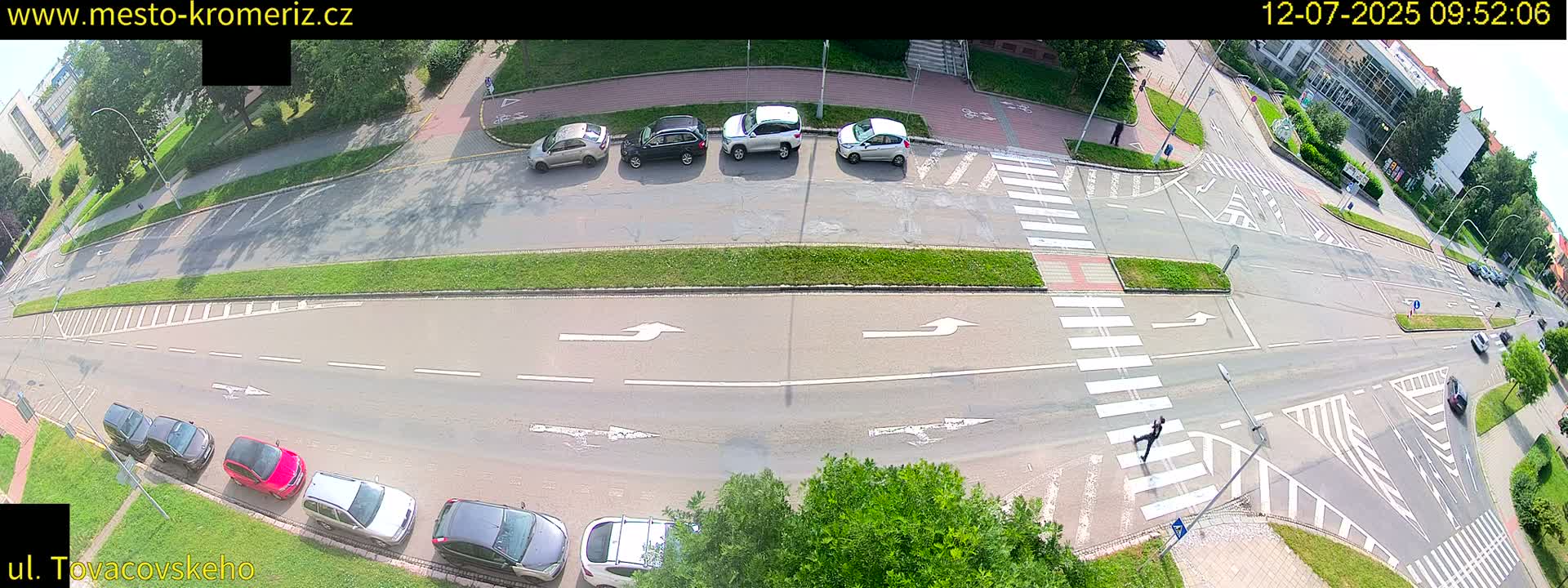 A sunny overhead view shows a street intersection with several cars parked and driving, and a pedestrian crossing a crosswalk.