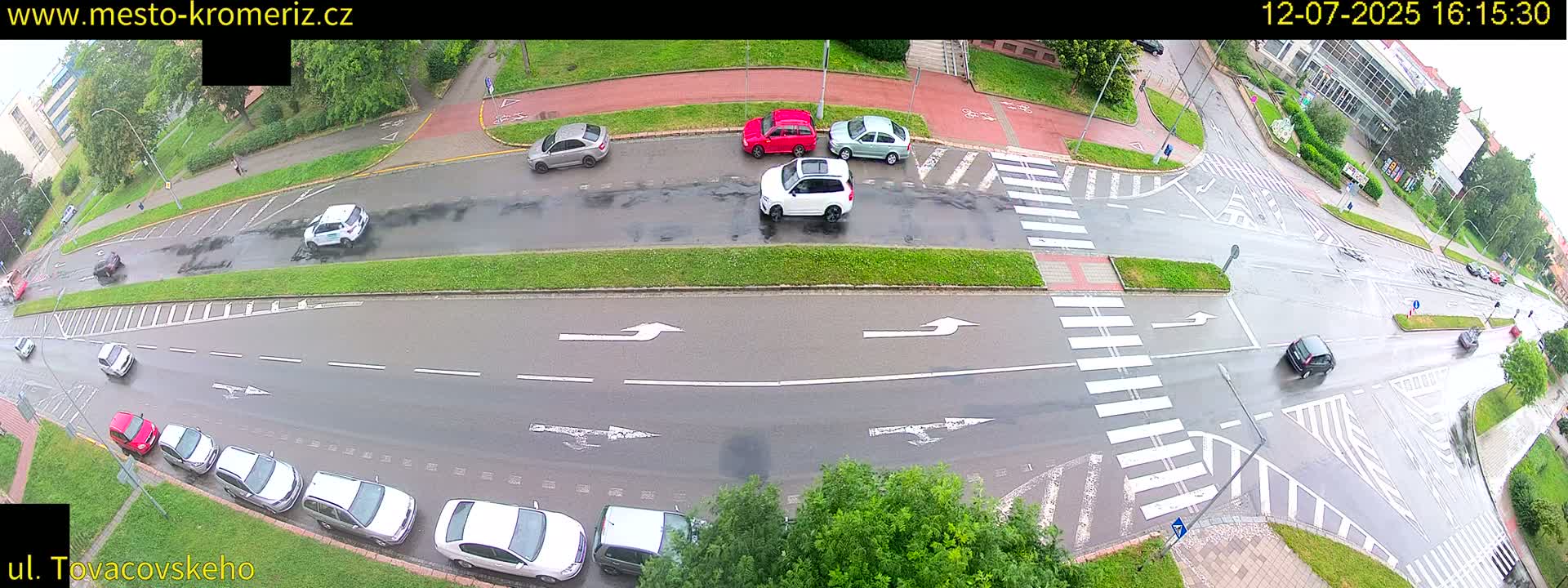 A sunny overhead view shows a street intersection with several cars parked and driving, and a pedestrian crossing a crosswalk.