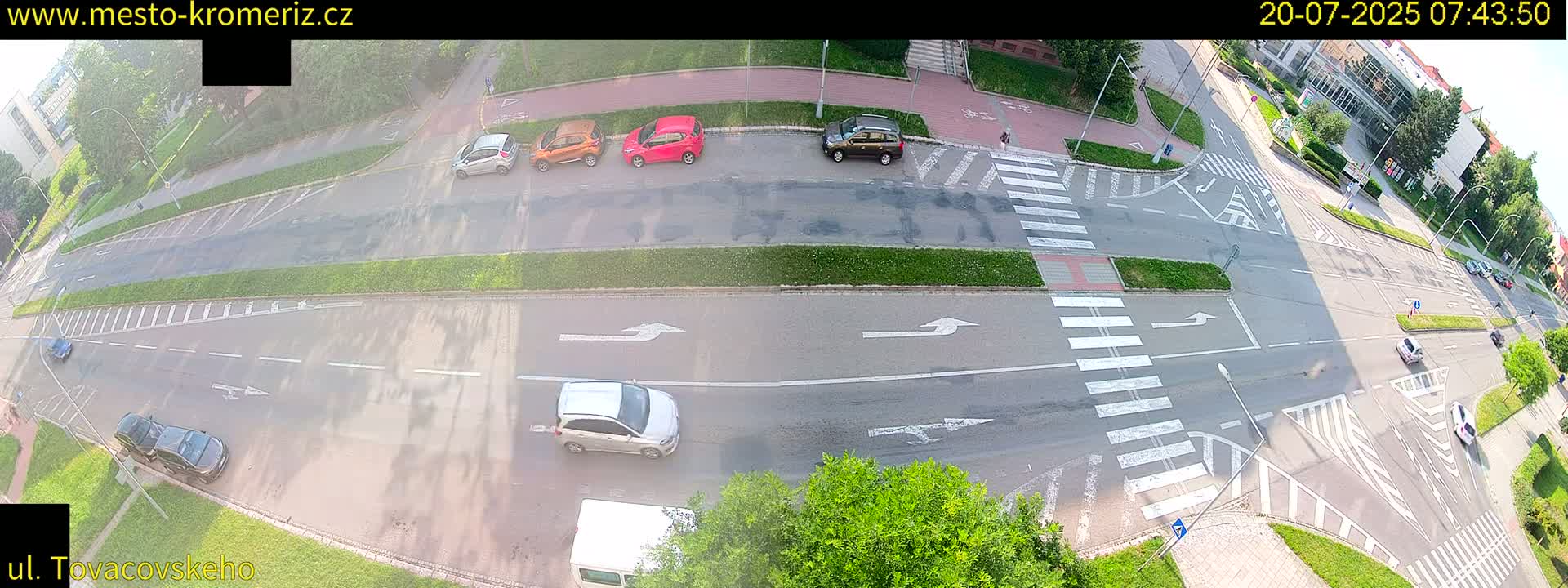 A sunny overhead view of a street intersection with several cars parked and driving, and pedestrians crossing a crosswalk.
