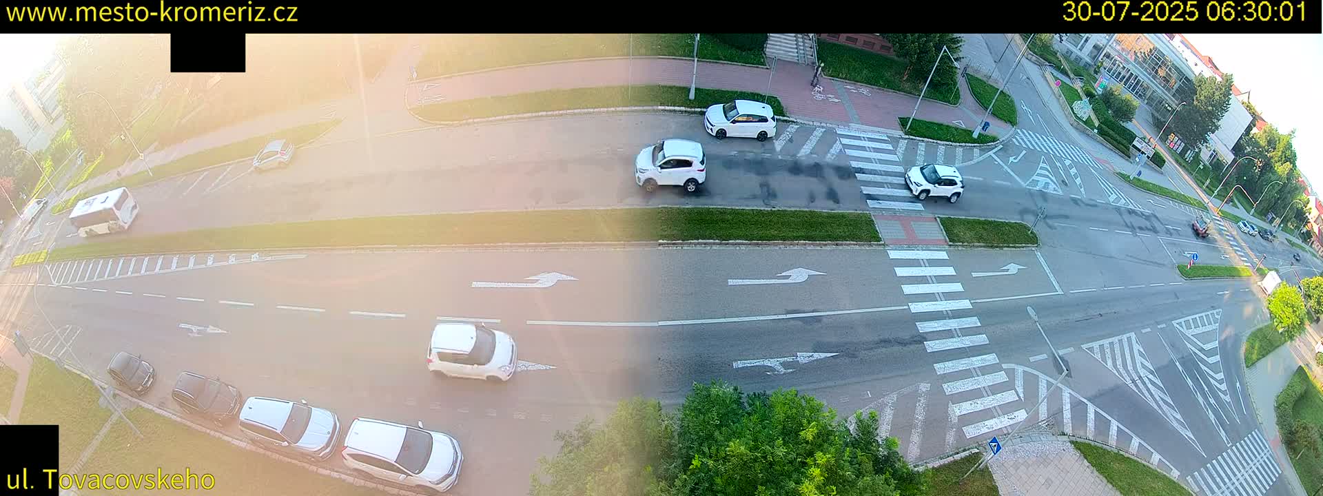 A sunny overhead view of a street intersection with several cars parked and driving, and pedestrians crossing a crosswalk.