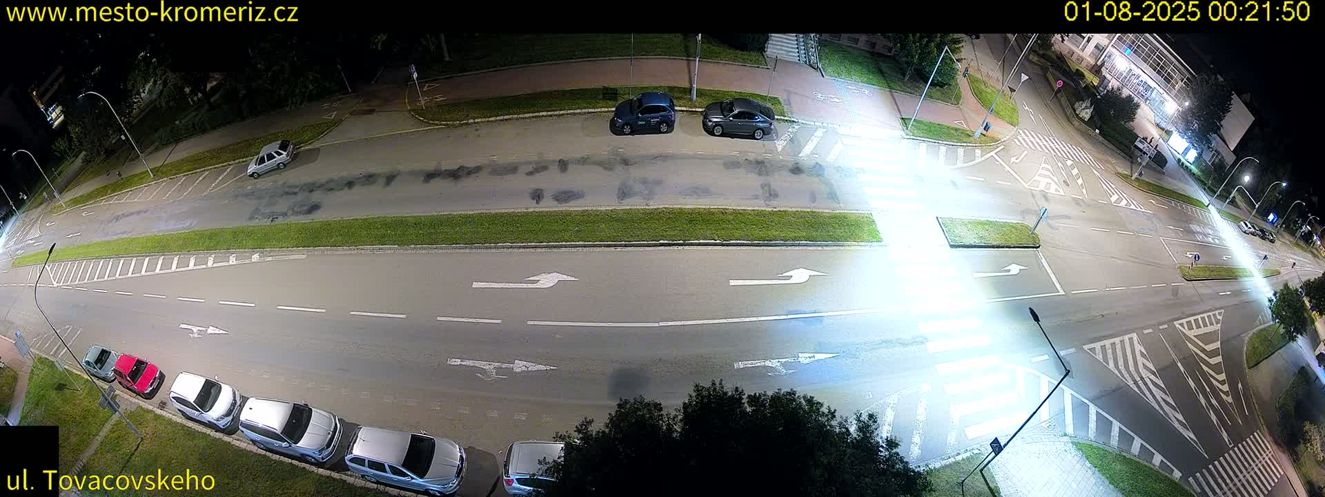 A nighttime overhead view of a street intersection with several parked cars and a few cars driving, under clear skies.