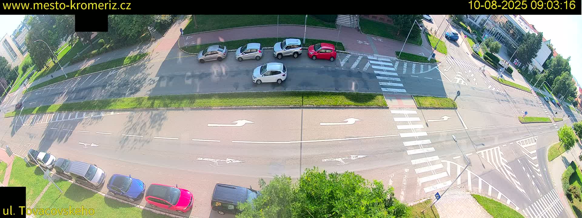 An overhead view shows a sunny day at a road intersection with several cars parked and driving, crosswalks, and bicycle lanes.