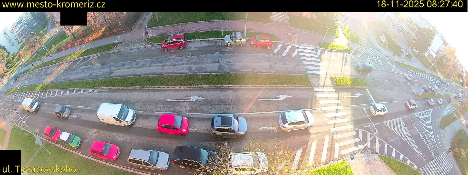 An overhead view shows a sunny day at a road intersection with several cars parked and driving, crosswalks, and bicycle lanes.