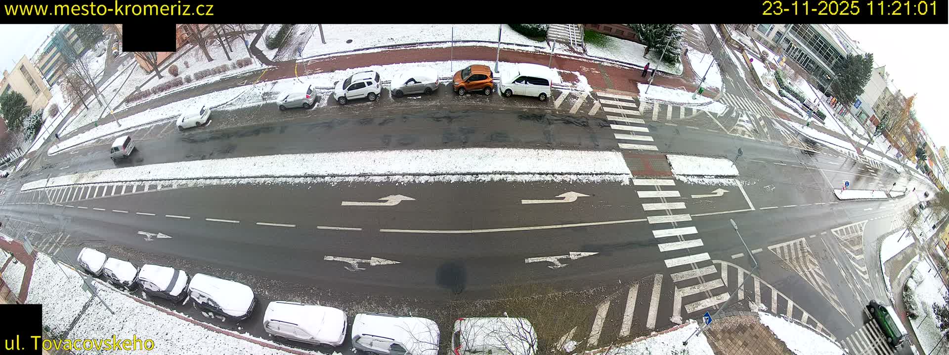 An aerial view captures a wide city street intersection and surrounding buildings on an overcast winter day, with patches of snow covering sidewalks and parked cars while the roads remain wet and mostly clear.