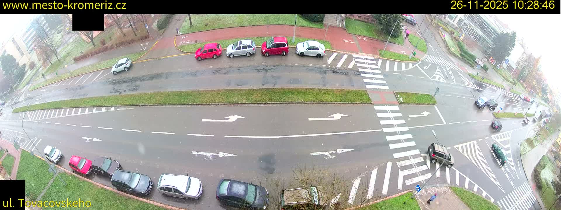 An elevated view captures a city street intersection with numerous parked cars and some moving vehicles on wet pavement, under an overcast sky with light precipitation.
