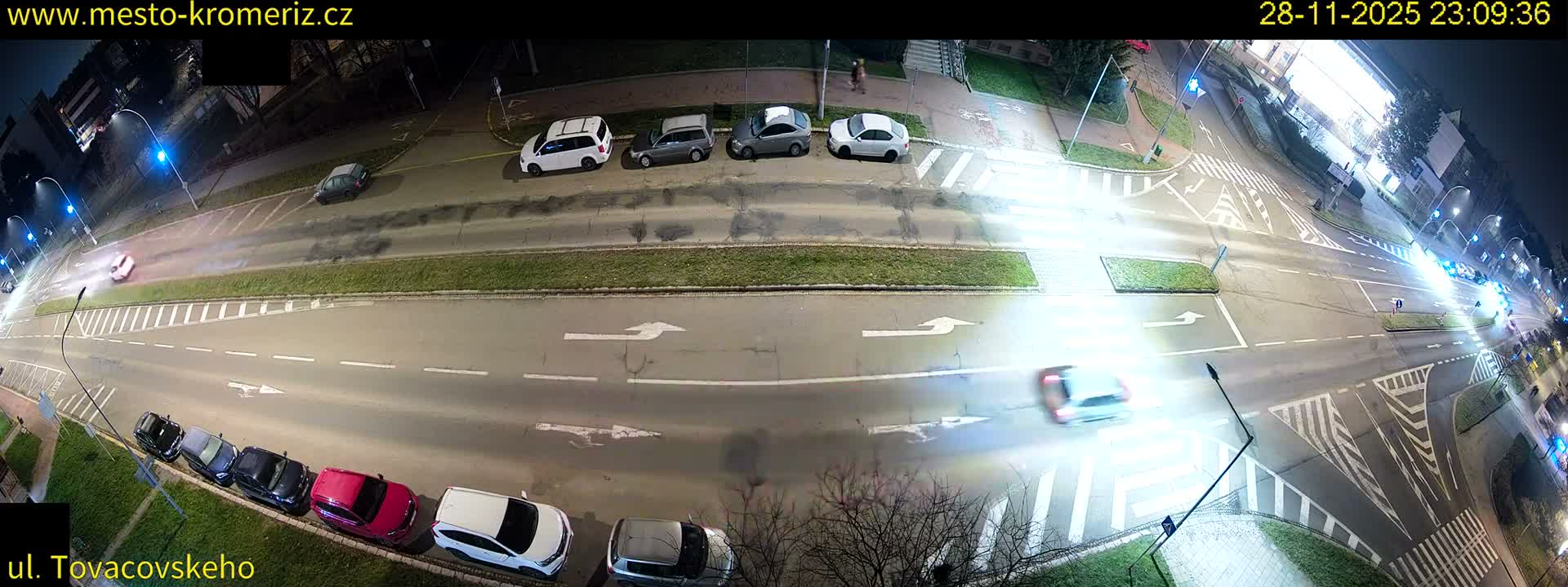 An elevated, clear weather, nighttime view captures a multi-lane city street and busy intersection, brightly illuminated by streetlights and vehicle headlights, with numerous cars parked along the side and others in motion.