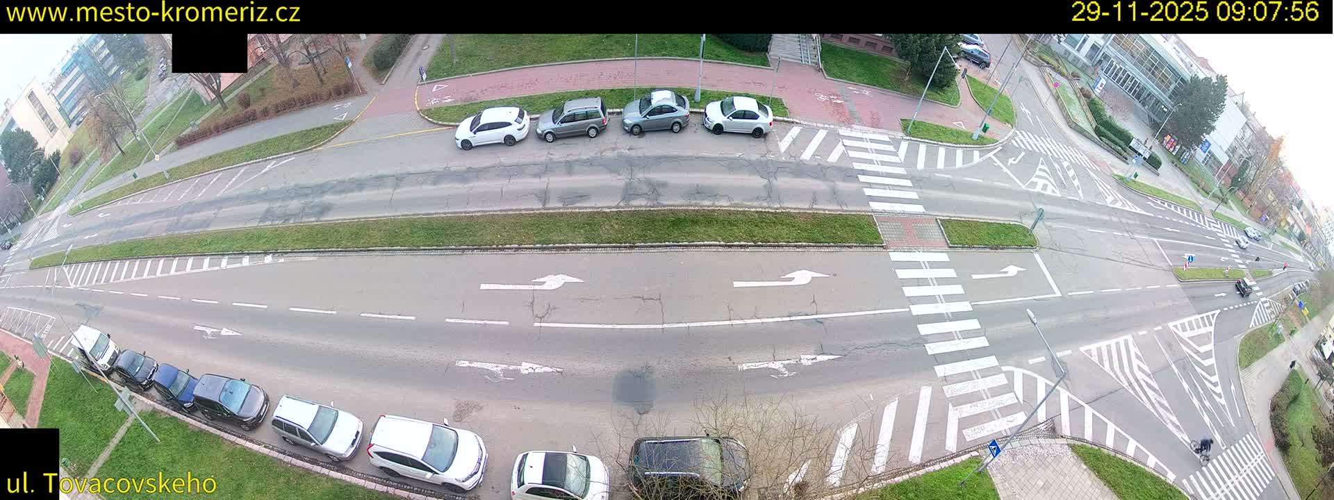 An elevated view captures an urban road intersection with multiple lanes, several parked and moving vehicles, pedestrian crossings, and grassy medians, under an overcast sky.