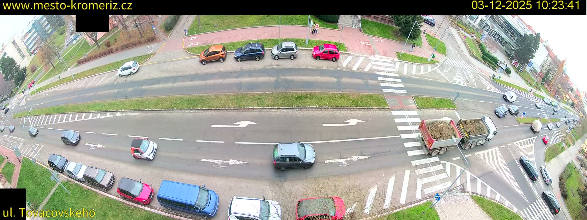 An aerial view captures a bustling urban intersection with numerous cars and a large dump truck navigating multi-lane roads, flanked by sidewalks with a few pedestrians and buildings in the distance, all under an overcast sky.