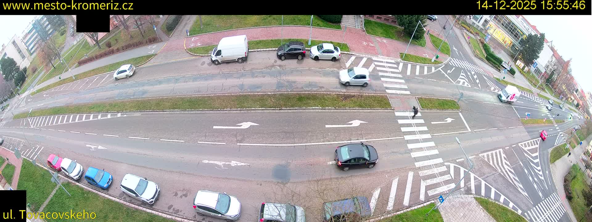 An aerial view captures a bustling urban intersection with numerous cars and a large dump truck navigating multi-lane roads, flanked by sidewalks with a few pedestrians and buildings in the distance, all under an overcast sky.