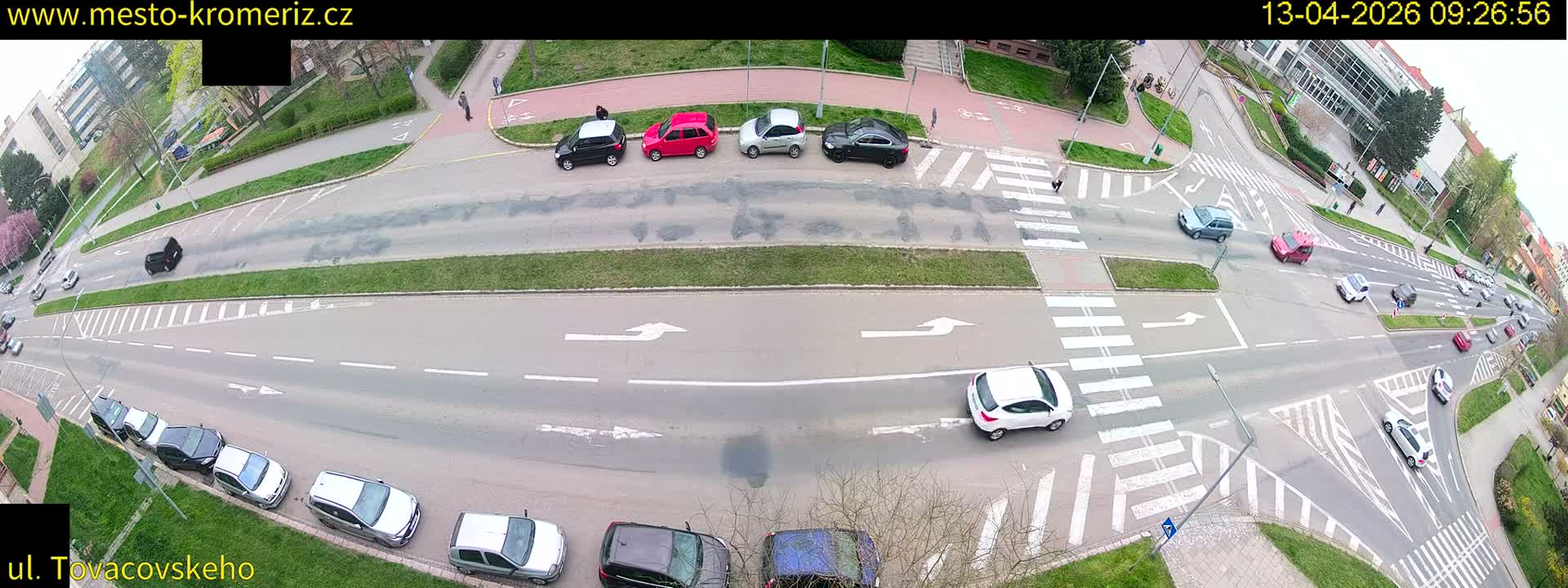 An aerial view captures a bustling urban intersection with numerous cars and a large dump truck navigating multi-lane roads, flanked by sidewalks with a few pedestrians and buildings in the distance, all under an overcast sky.