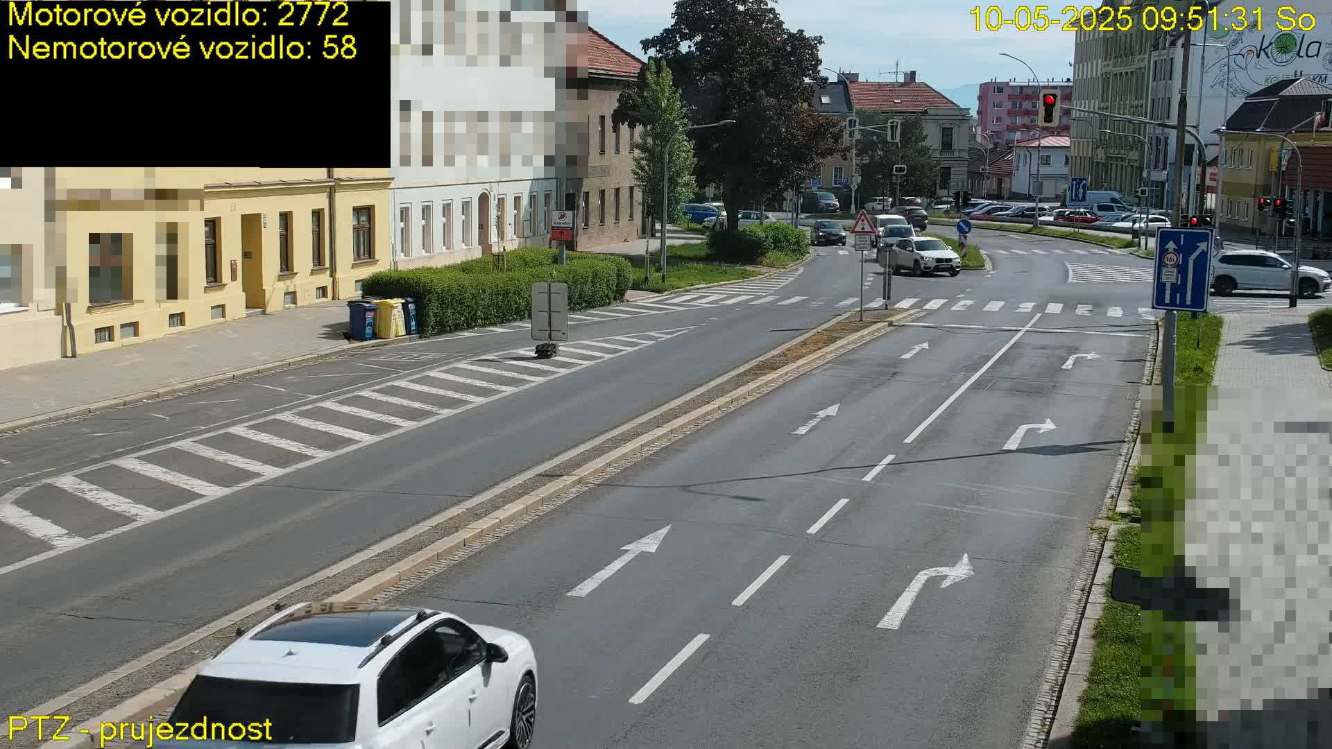 A sunny day shows a multi-lane road intersection with several cars, buildings, and pedestrian crosswalks.
