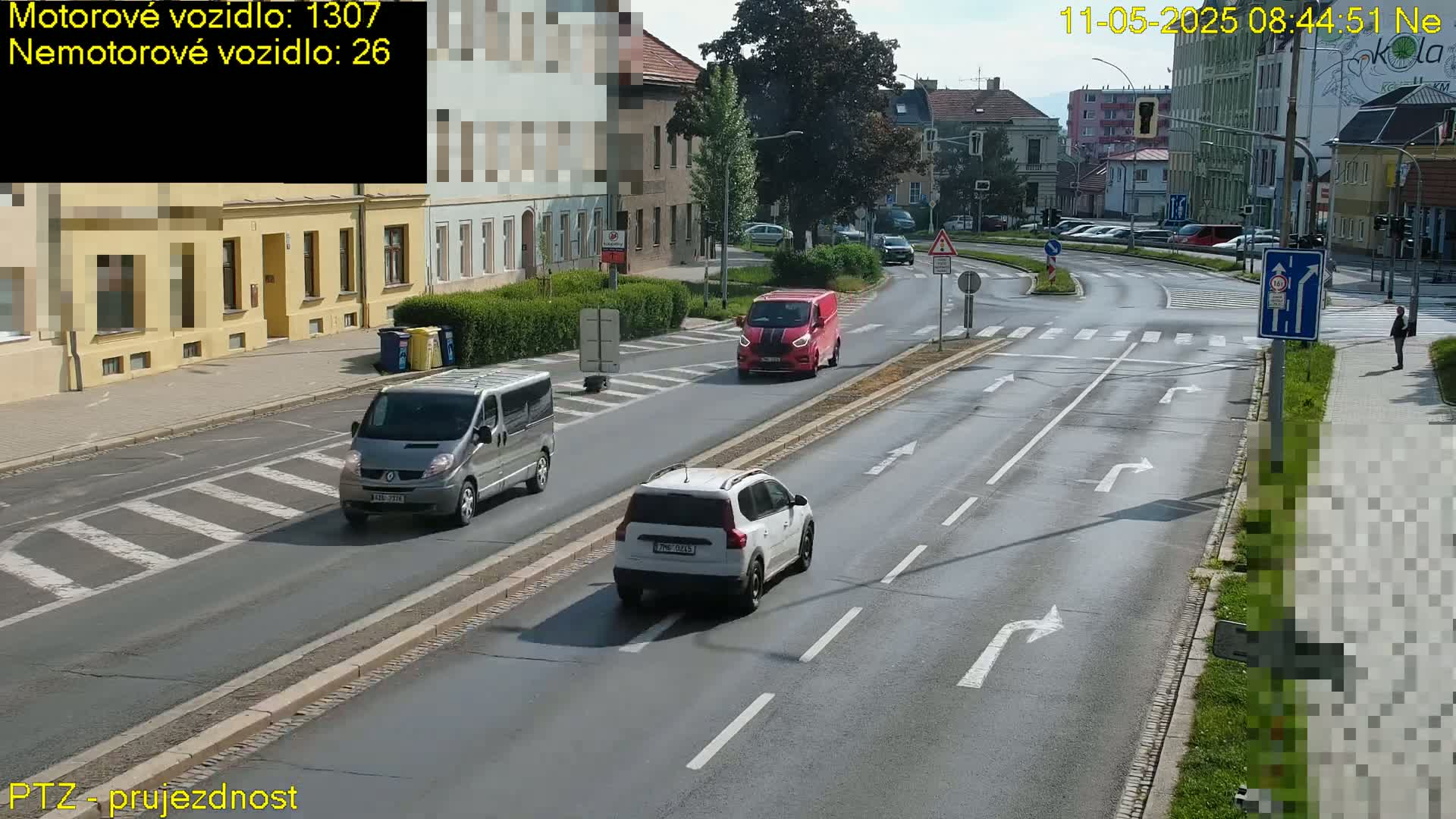 A sunny day shows a multi-lane road intersection with several vehicles, including a red van and a white SUV, and a pedestrian walking on the sidewalk.