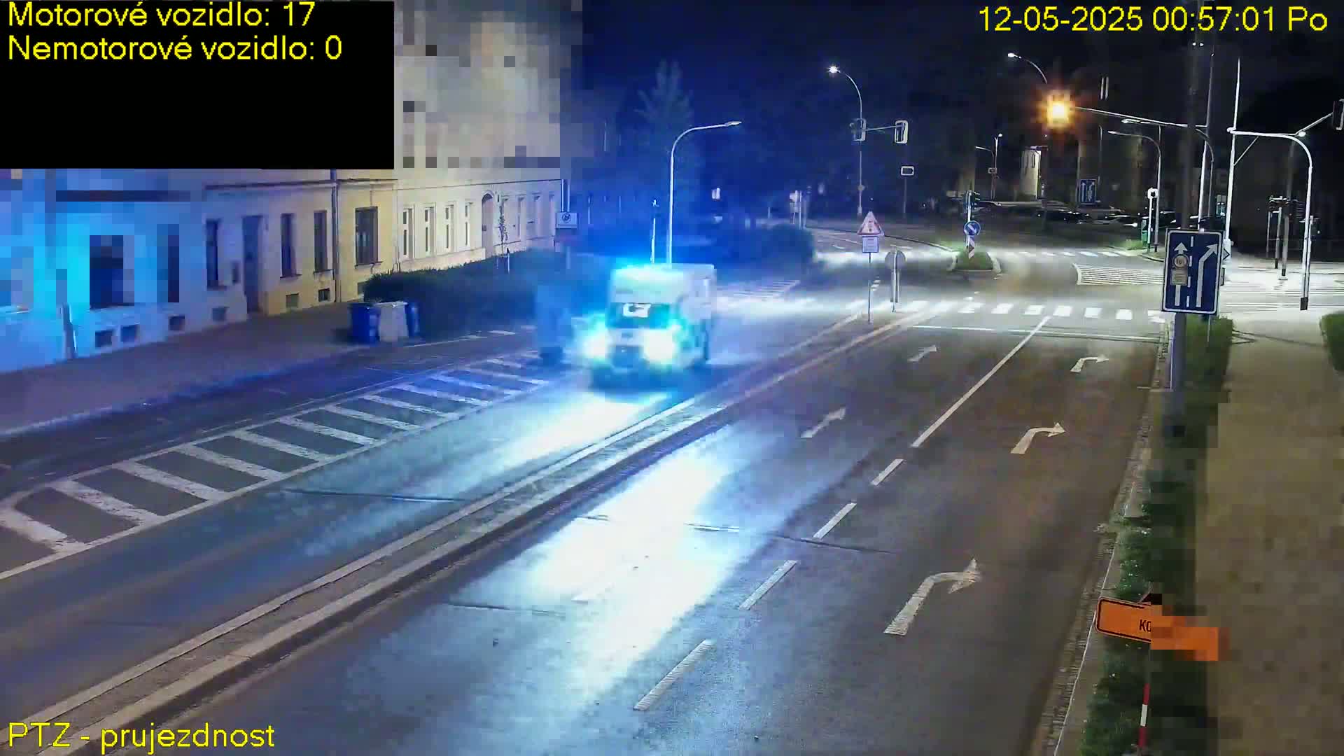 A white van with flashing lights drives down a dark, wet street at night, passing a crosswalk and streetlights.