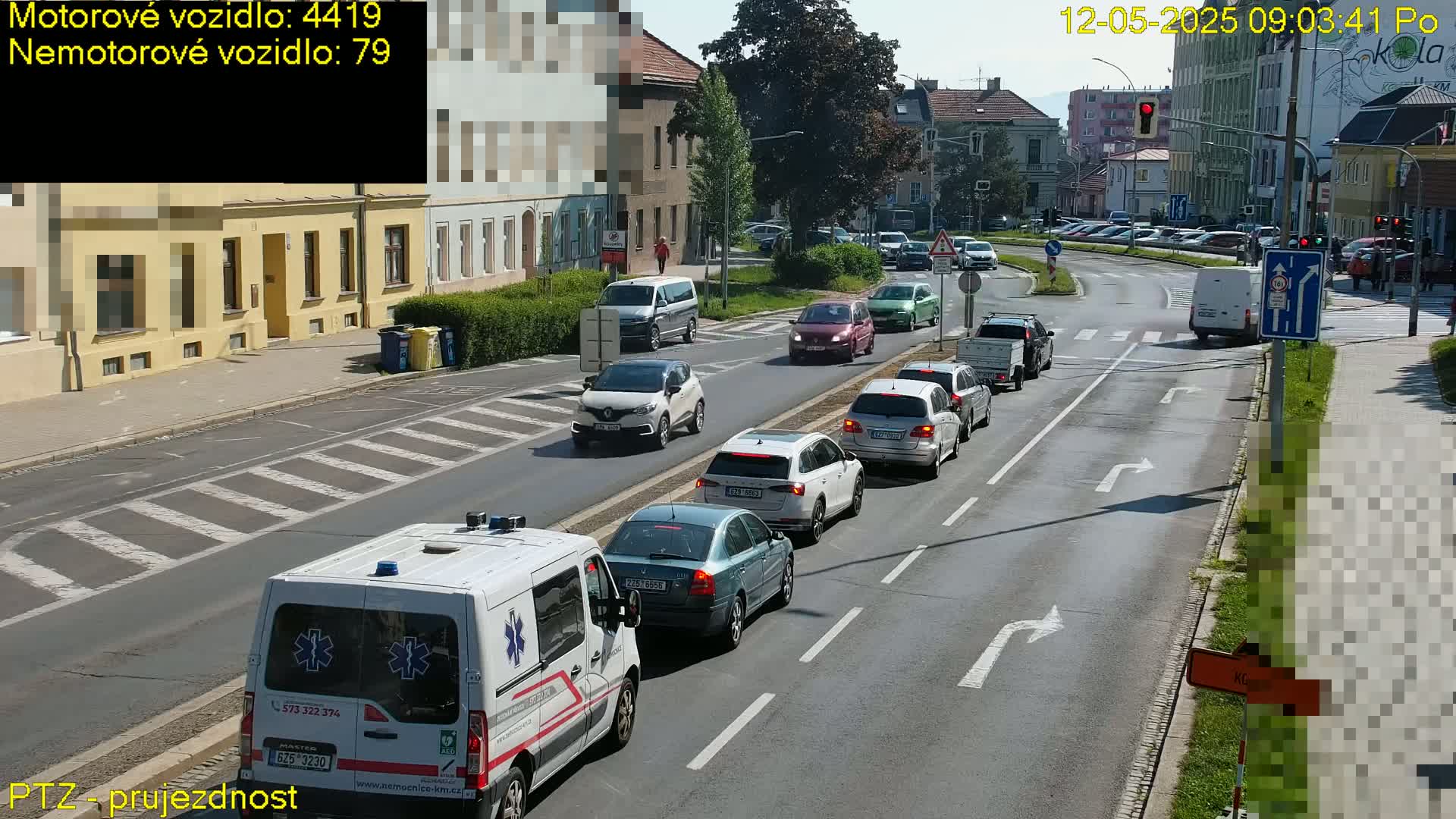 A sunny day shows several cars and a van stopped at an intersection, with an ambulance at the front of a line of vehicles.