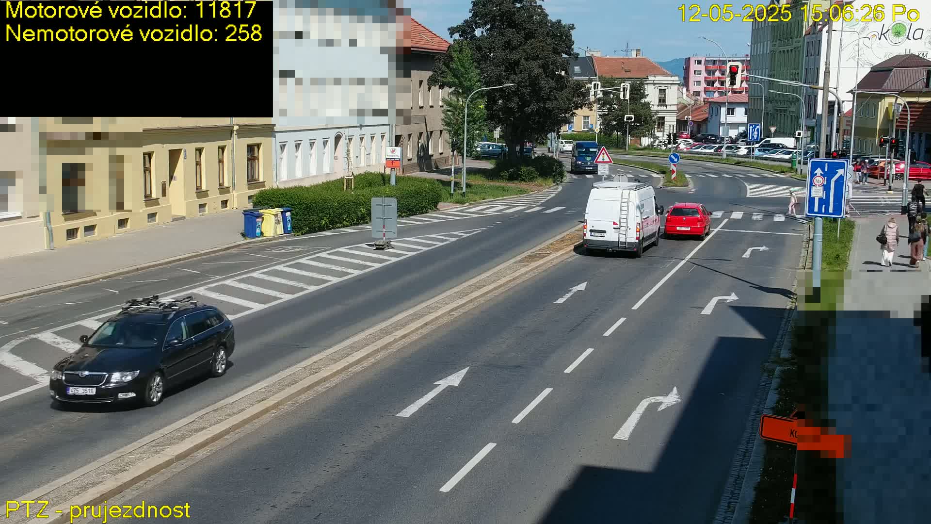 A sunny day shows a multi-lane road with several cars, a van, and pedestrians crossing at a crosswalk.