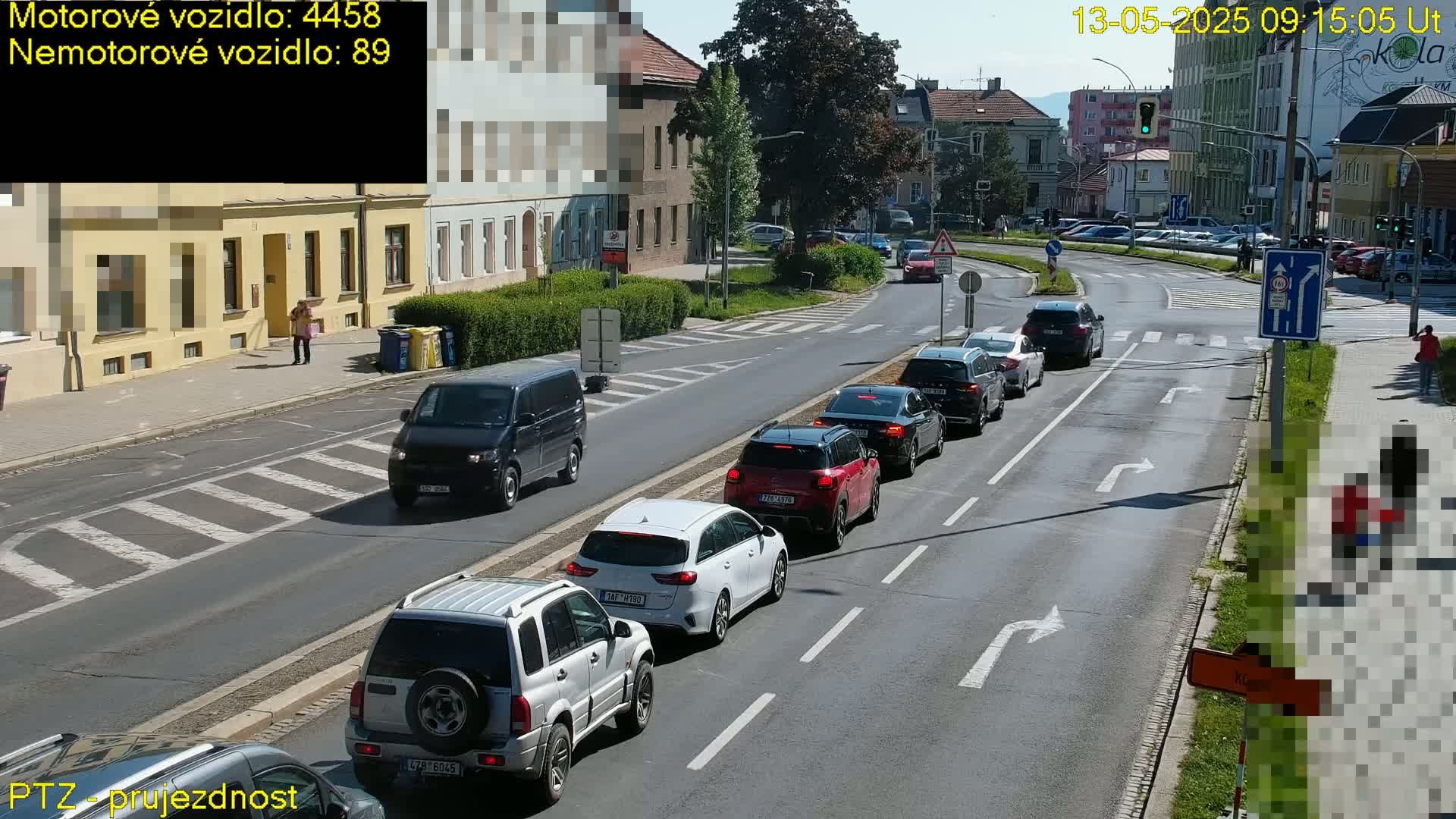 On a sunny day, several cars are stopped at an intersection, with a few pedestrians visible on the sidewalk.