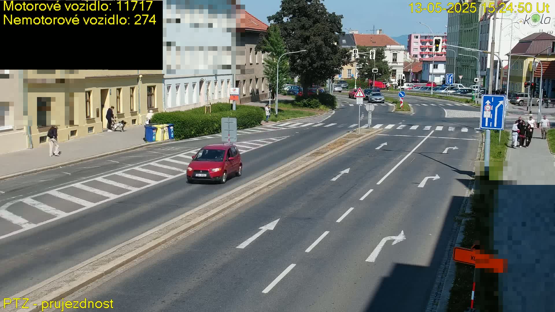 A sunny day shows a red car driving on a multi-lane road with pedestrians and dogs on the sidewalk, and buildings lining the street.