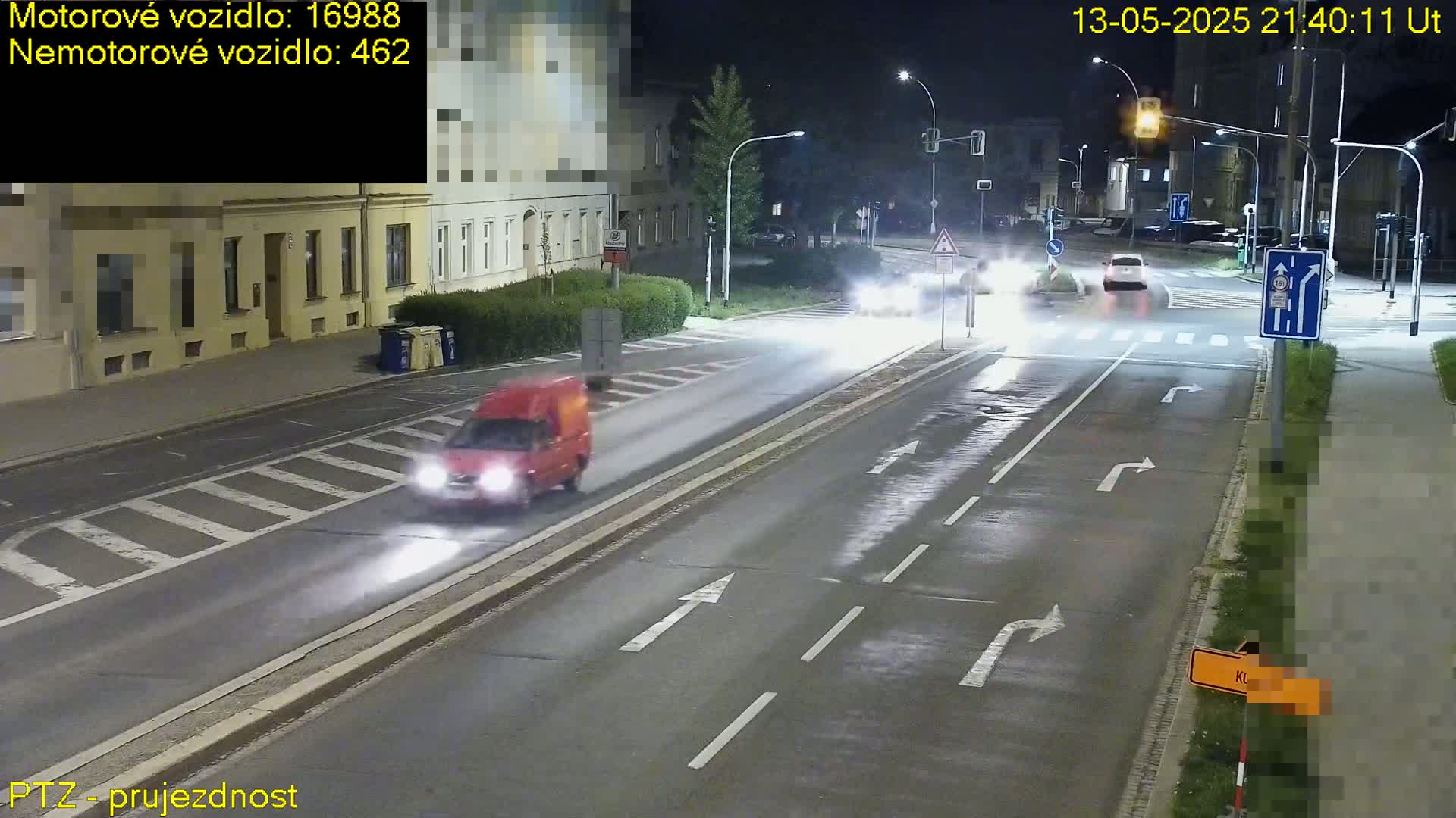 A red van drives on a wet road at night past a crossroads with other vehicles in the distance under streetlights.