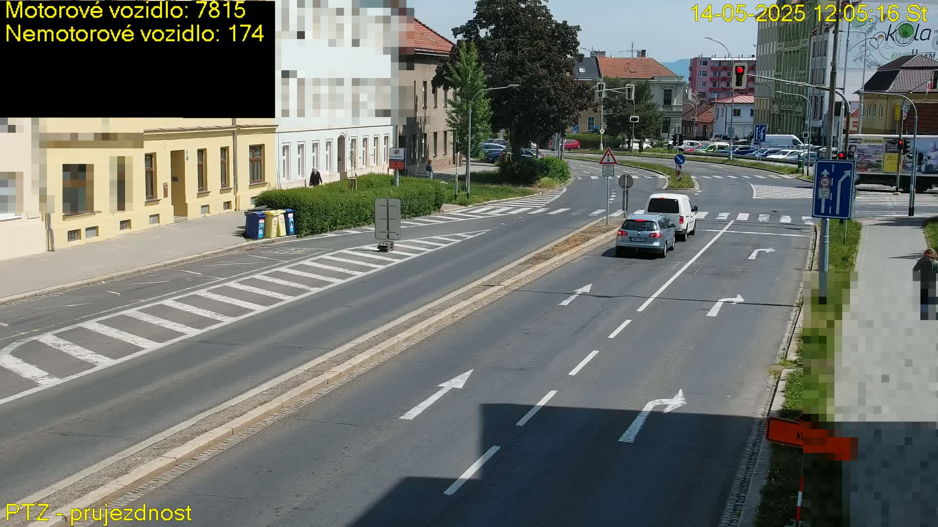 A sunny day shows a multi-lane road with several cars at an intersection near buildings, a pedestrian crossing, and some pedestrians.