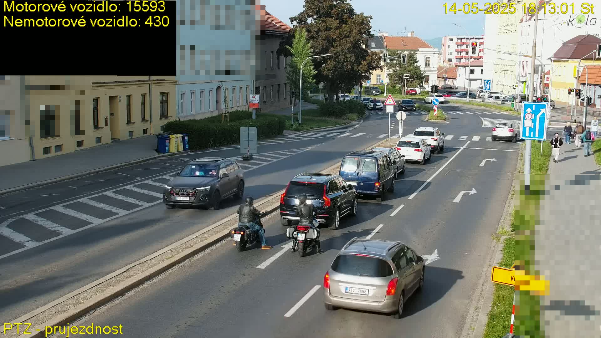 A sunny day on a city street shows several cars and two motorcycles stopped at an intersection with pedestrians crossing.